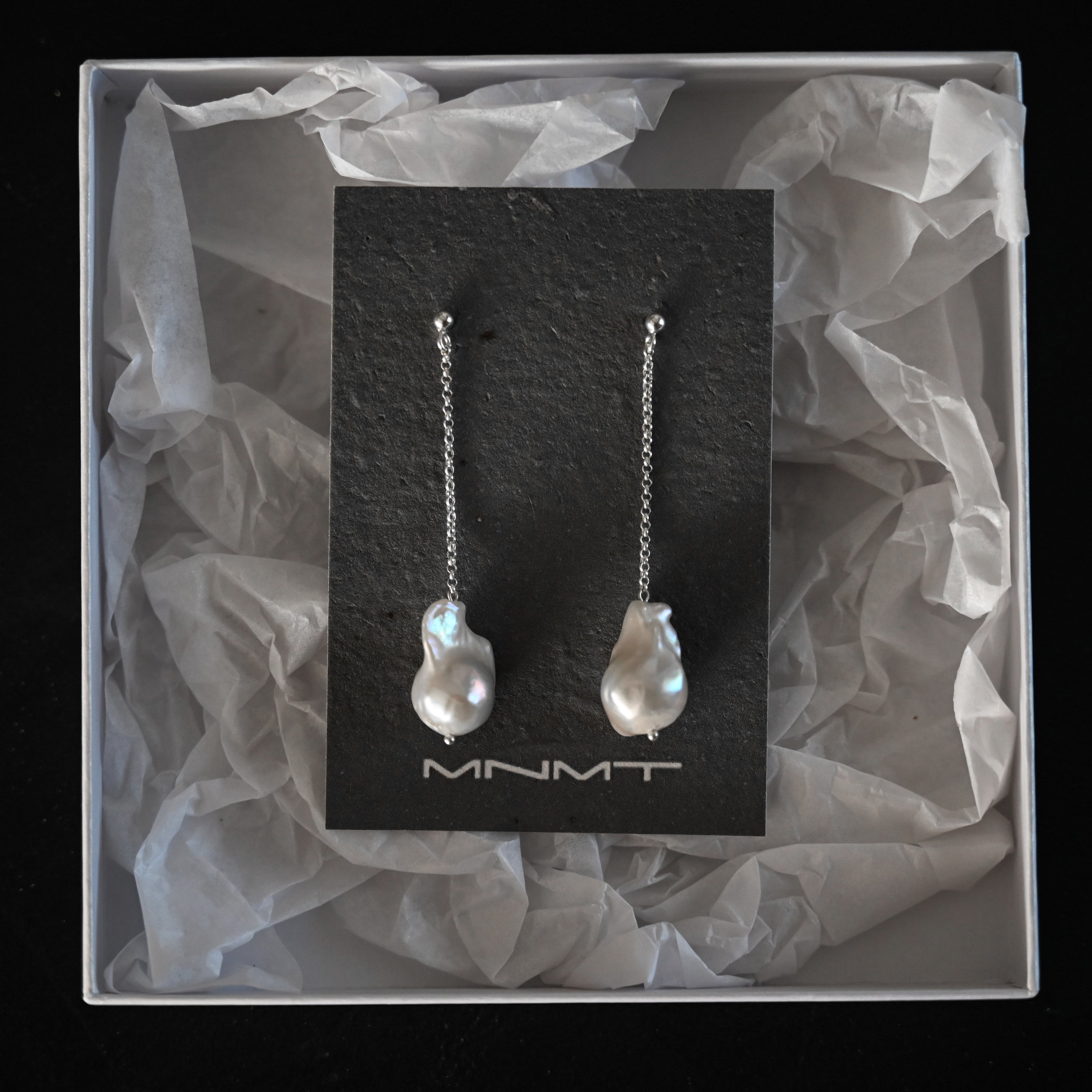 Pair of pearl earrings on a black card with 'MNMT' branding, inside a box with crumpled tissue paper.