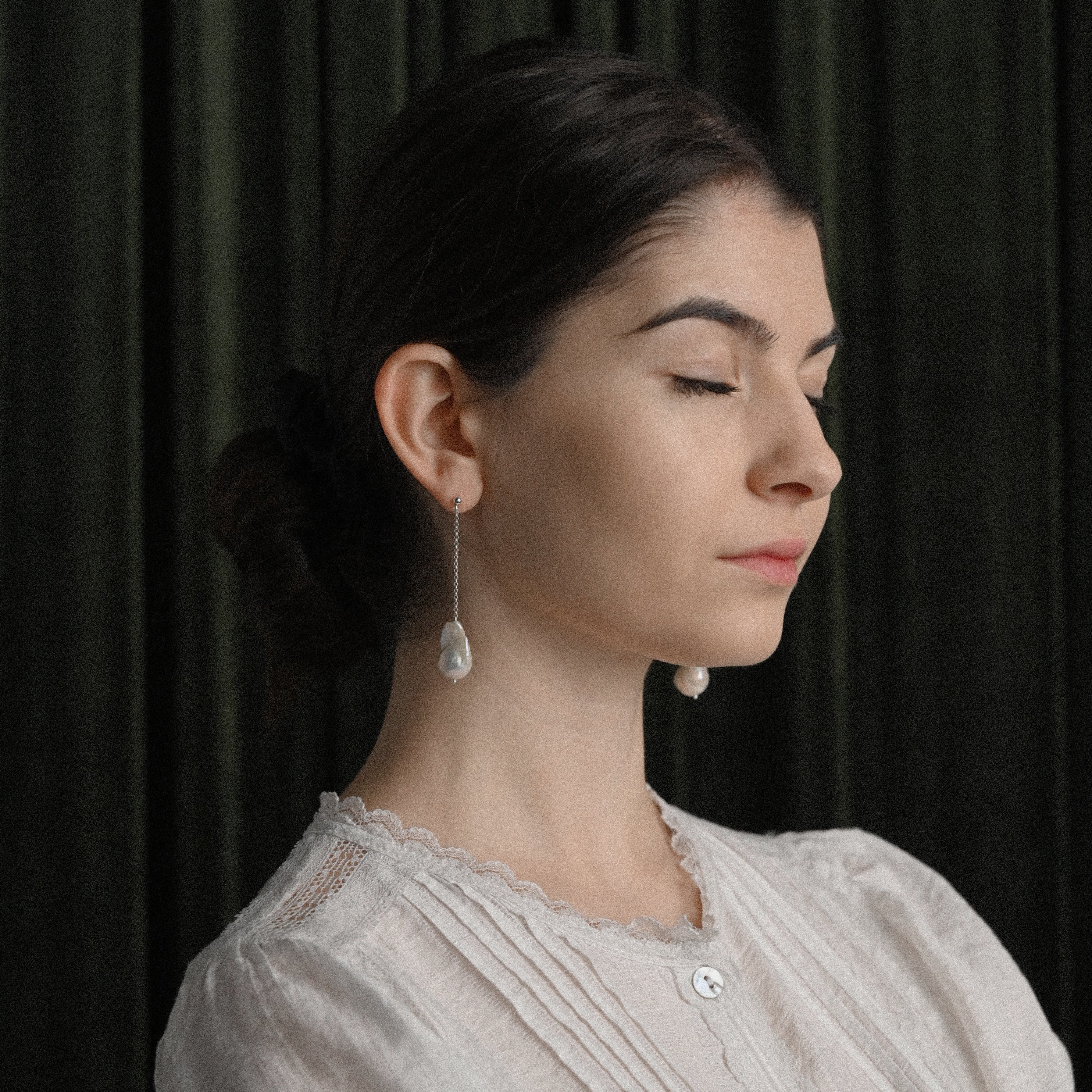 Woman wearing long white pearl earrings against a dark curtain background