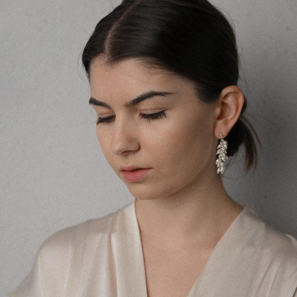 Woman wearing a beige top and white pearl silver earrings against a plain background