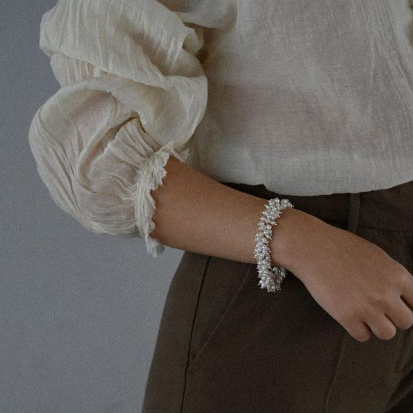 Close-up of a woman wearing a white blouse with ruffled sleeves and whiet pearl bracelet against a gray background.