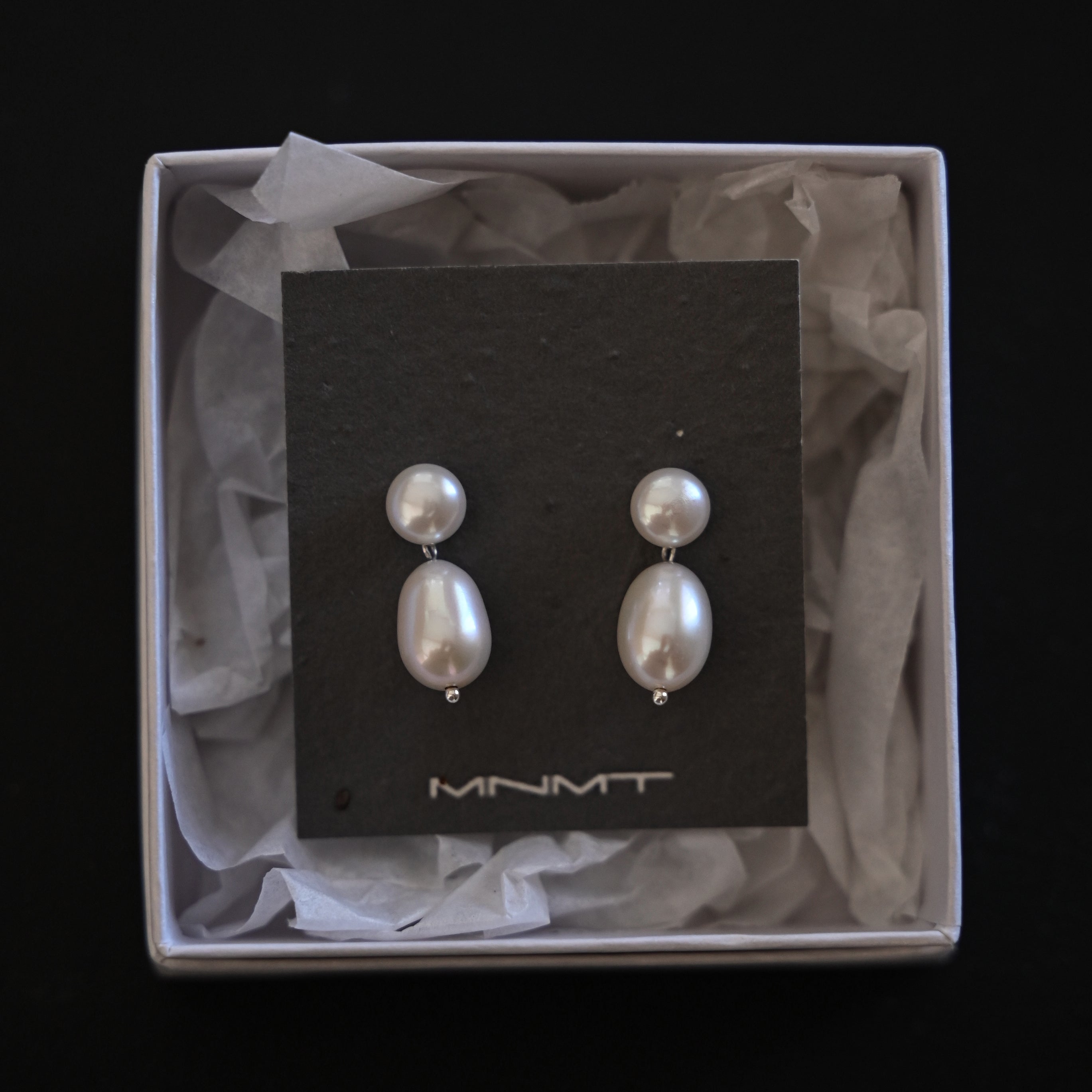Pair of pearl earrings on a black card with 'MNMT' branding, displayed in a box.