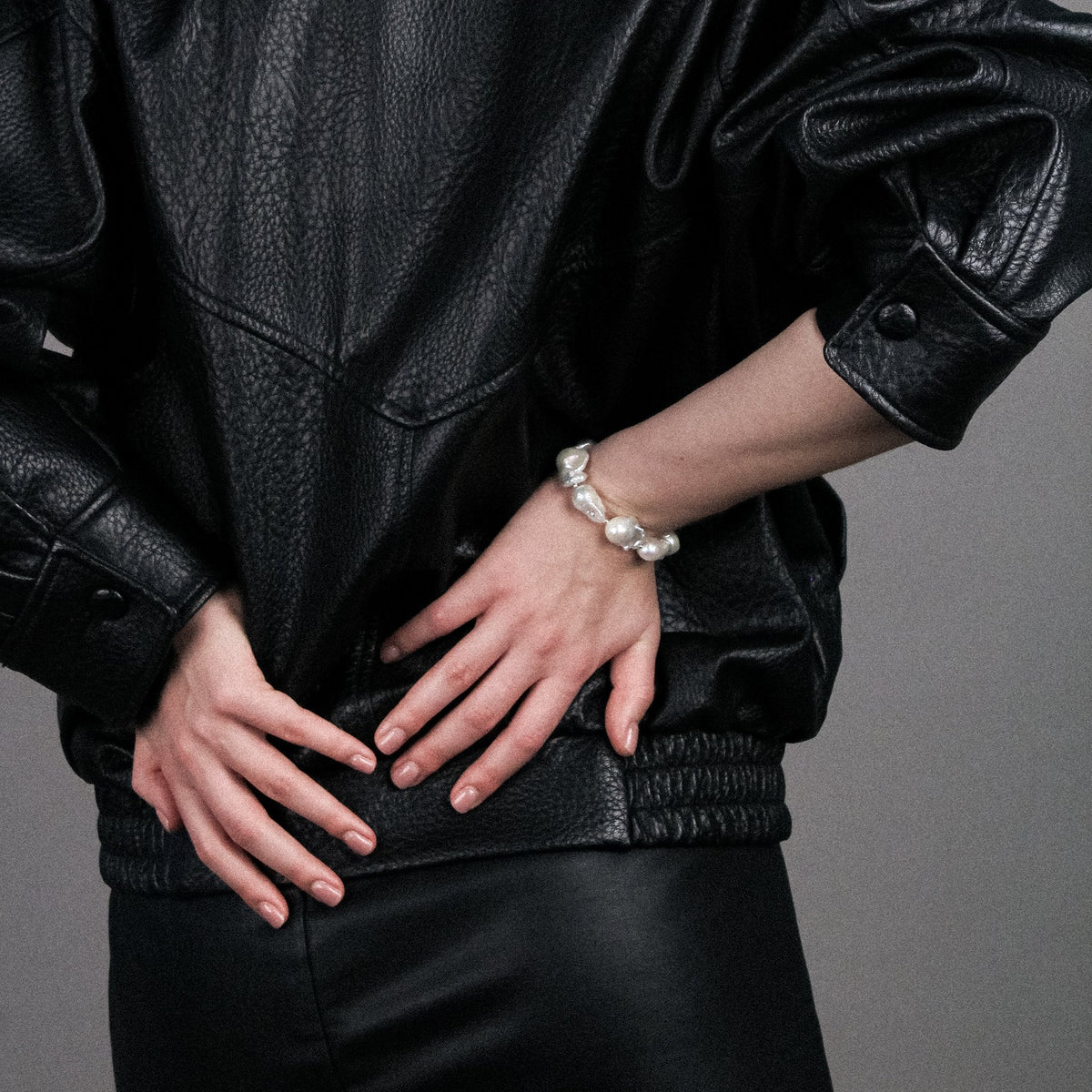 A close-up of a person wearing a black textured leather jacket and black pants, with one hand resting on the hip and the other showing a white pearl bracelet, against a solid gray background.